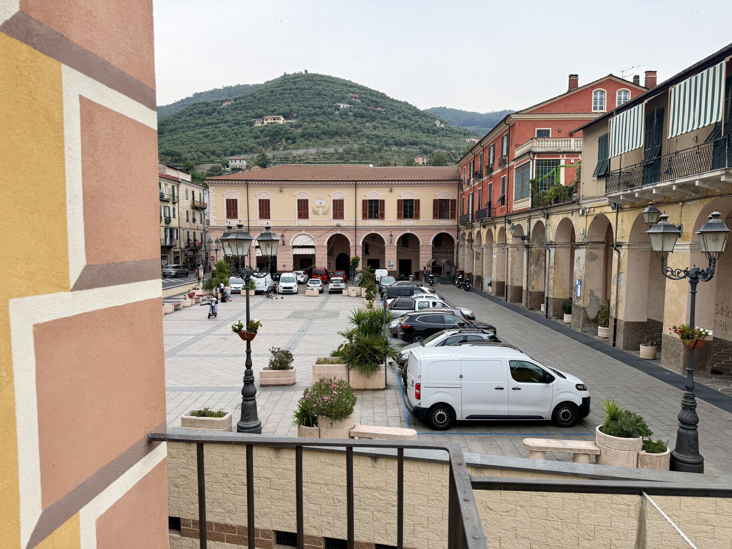 Pátio com carros estacionados, uma van branca e um edifício com arcos, com uma montanha ao fundo.