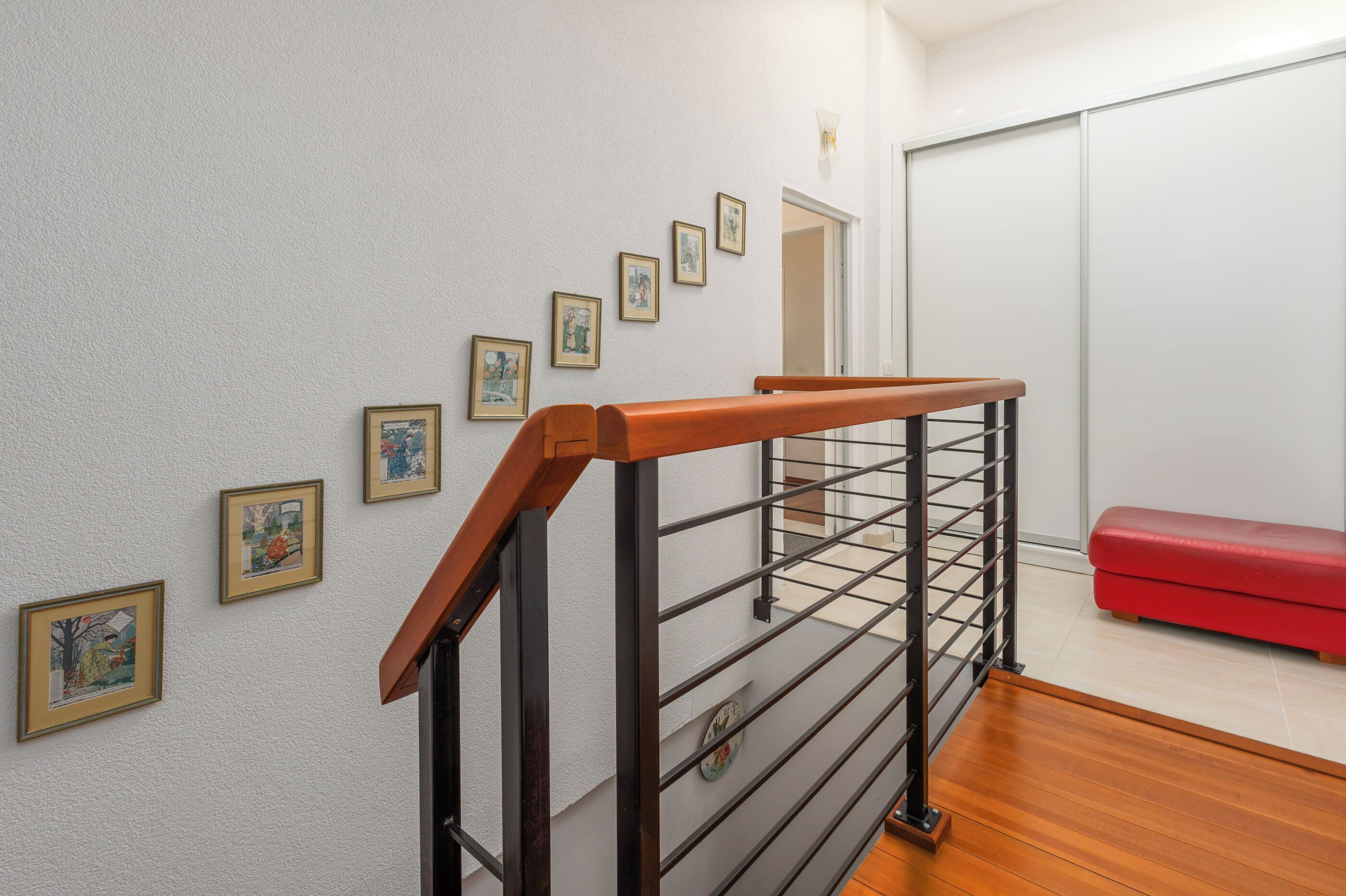A staircase with a wooden handrail and metal railing, a red bench, and framed pictures on the wall.