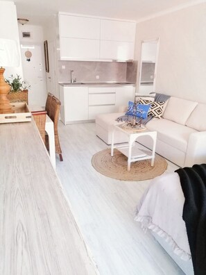 Ein modernes Wohnzimmer mit Essbereich, einer Küche mit weißen Schränken und einem weißen Sofa mit gemusterten Kissen.
