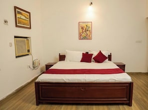 A bedroom with a wooden bed, white bedding, red pillows, a red throw, a wall-mounted air conditioner, a framed picture, and a wall-mounted light fixture.