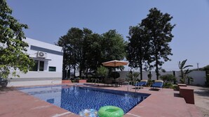 Une piscine entourée d’arbres et d’un bâtiment, avec des chaises longues et un parasol.