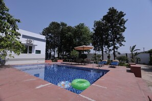 En swimmingpool med solstolar och ett parasoll, omgiven av trÀd och en byggnad.