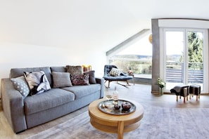 Ein modernes Wohnzimmer mit einem grauen Sofa, einem runden Couchtisch aus Holz und einem blauen Sessel.