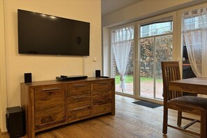 Een kamer met een wandgemonteerde flatscreen televisie, een houten kast met lades, een eettafel met een stoel, en een schuifdeur van glas die leidt naar een buitenruimte.