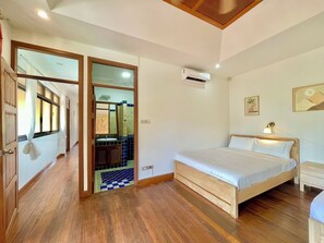 Ein Schlafzimmer mit einem hölzernen Bett, einer Klimaanlage, einer Nachttischlampe und einem Badezimmer mit gefliestem Boden und einem Spiegel.