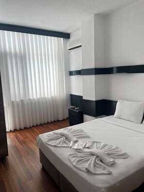 A hotel room with a neatly made bed, a bedside table with a remote control, and a window with sheer curtains.