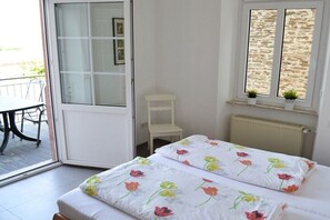Ein Schlafzimmer mit Bett, einem Sessel, einem Fenster mit Pflanzen und einer Tür, die auf einen Balkon führt.