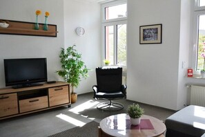 Un salon moderne avec un téléviseur à écran plat, un meuble en bois, un fauteuil noir, une table ronde agrémentée d’une plante et une fenêtre laissant entrer la lumière naturelle.