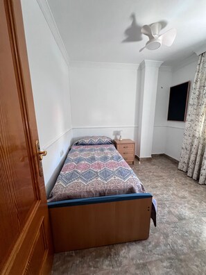 Un dormitorio con una cama individual, una mesita de noche, un ventilador de techo y una ventana con cortinas.