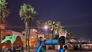 A beach with a playground, palm trees, and buildings in the background. - Apartment Sleeps 7 With Balcony (Velez-Malaga)