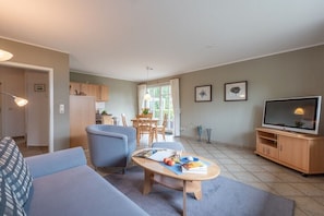 Ein Wohnzimmer mit einer Couch, einem Couchtisch mit Büchern und Früchten, einem Fernseher und einem Essbereich mit Tisch und Stühlen.