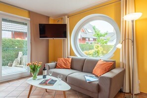 Ein Wohnzimmer mit einer Couch, einem Rundfenster, einem Flachbildfernseher und einem Tisch mit Blumen und einer Weinflasche.
