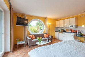 Ein kompaktes Hotelzimmer mit einem Bett, einer Couch, einem kleinen Tisch mit einer Vase, einer Küchenzeile mit Schränken und Geräten sowie einem großen Fenster.