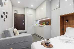 Ein modernes Hotelzimmer mit grauem Sofa, einem Bett mit weißer Bettwäsche, einer Küchenzeile und einer Holztür.