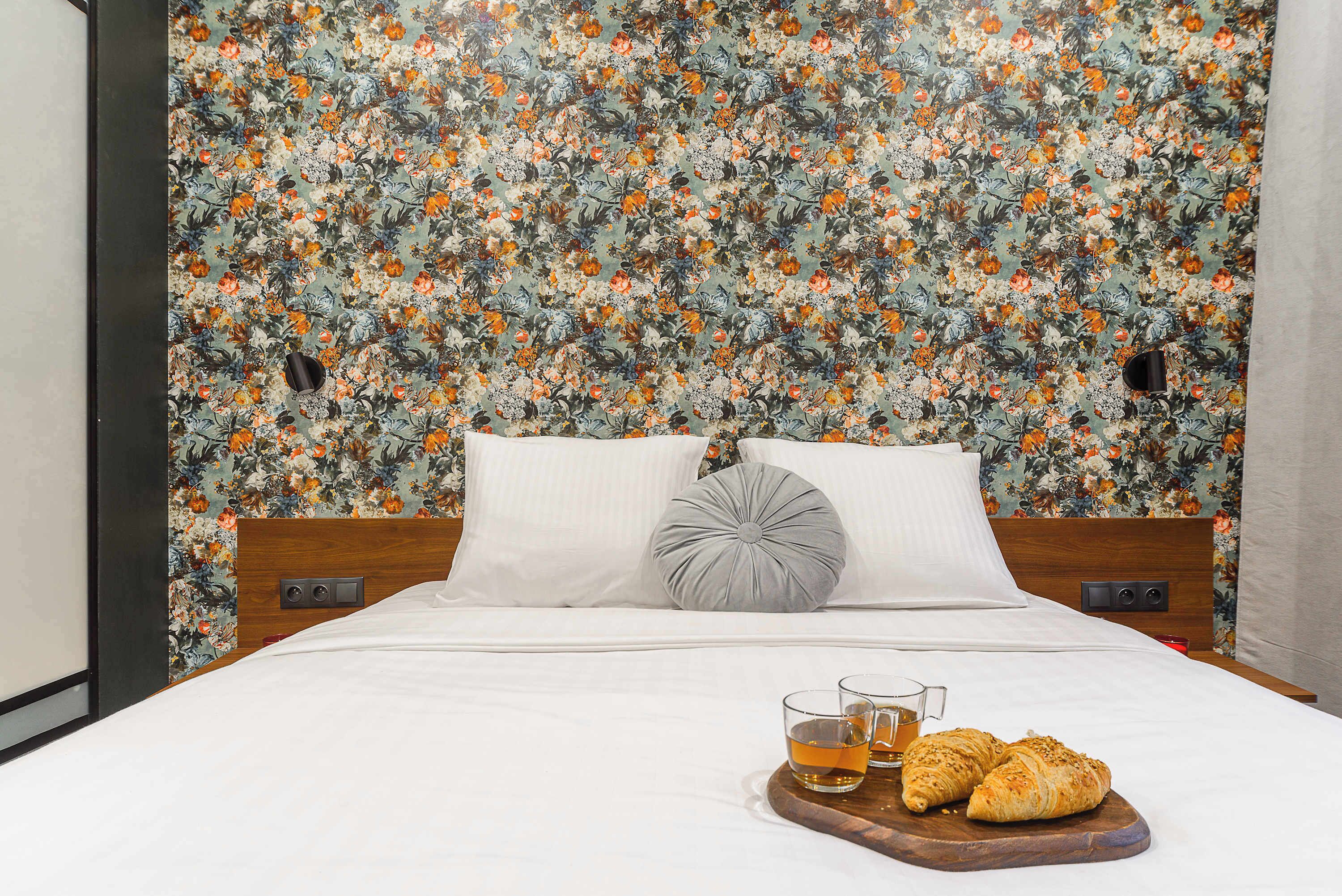 A hotel room with a floral wallpaper headboard, a bed with white linens, two pillows, a bedside table with a glass of tea and croissants, and a wooden headboard.