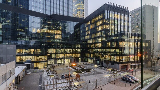 Een modern gebouw met meerdere glazen gevels, een binnentuin met zitplaatsen en een parkeerplaats.