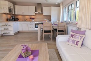 Eine moderne Küche mit weißen Schränken, ein Essbereich mit Tisch und Stühlen sowie ein Wohnzimmer mit einer Couch und einem Zierkissen.