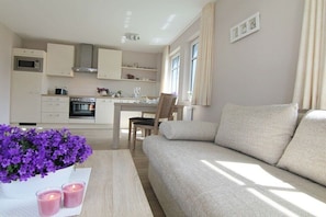 Eine moderne Küche mit weißen Schränken, ein Essbereich mit Tisch und Stühlen sowie ein Wohnzimmer mit beigefarbenem Sofa.
