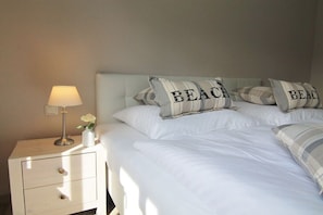 Ein Bett mit weißer Bettwäsche, zwei Kissen mit dem Aufdruck 'BEACH', eine Nachttischlampe und ein kleiner Blumenstrauß in einer Vase.