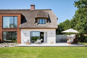 Ein modernes Haus mit einer großen Glasdoor, einer Terrasse mit Gartenmöbeln und einem Strohdach.