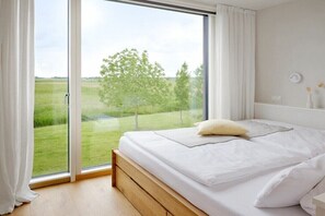 Ein Schlafzimmer mit einem großen Schiebeglasfenster, das zu einer Rasenfläche führt, einem Bett mit weißer Bettwäsche und einem Nachttisch mit einer Lampe.