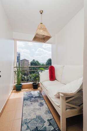Ein Balkon mit einer weißen Couch, einem roten Kissen, Blumentöpfen und einer gewebten Deckenleuchte.