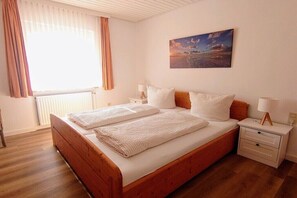 Ein Schlafzimmer mit einem hölzernen Bett, zwei Nachttischen, einem Schminktisch, einem Fenster mit Vorhängen und einem Bild an der Wand.