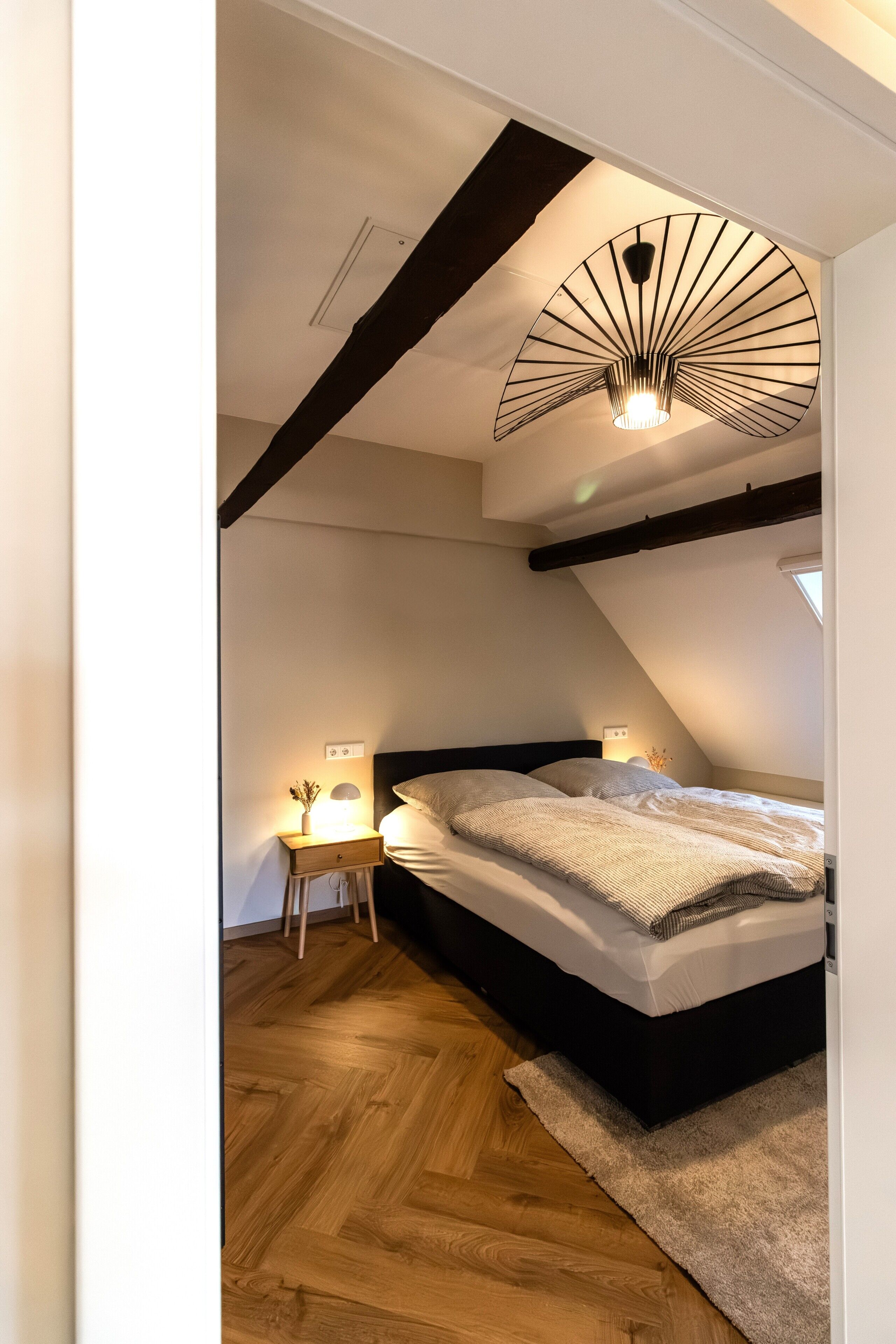 Une chambre avec un plafond en pente, un lit avec une tête de lit, une table de chevet avec une lampe et un plancher en bois.