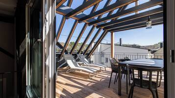 Eine Holzterrasse mit zwei Liegestühlen und einem Tisch, eine Glastür, die zu einem Außenbereich führt, und ein klarer Himmel.