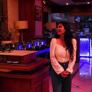 A woman standing in a bar area with a counter displaying glasses and a plant. - Hotel Supreme Heritage (Pune)