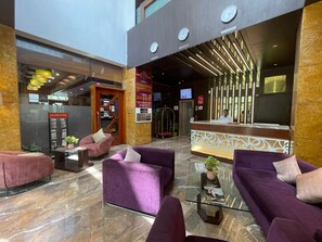 A hotel lobby with purple seating, a reception desk, clocks, and a Times of India display. - Hotel Supreme Heritage (Pune)