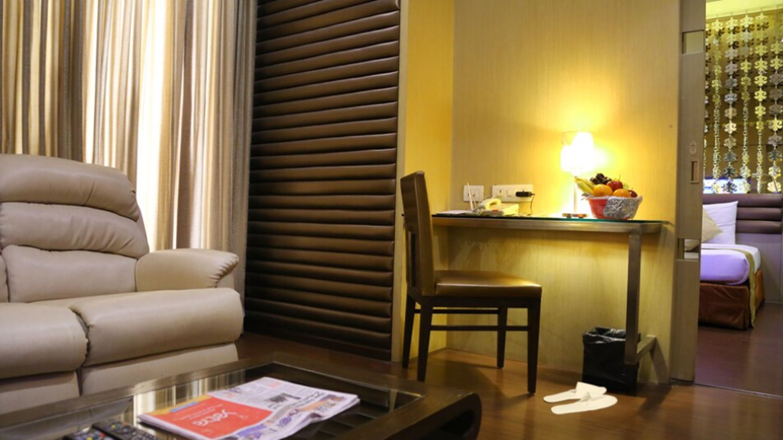 A hotel room with a sofa, a dining table with a lamp, a chair, and a small trash bin.