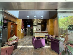 Eine Lobby mit einer Rezeption, violetten Sofas, einem Glastisch und einem großen Fenster mit Blick auf Bäume.