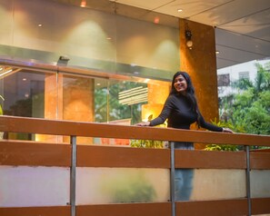 A person standing on a balcony with a railing, overlooking a building with glass windows and a view of greenery. - Hotel Supreme Heritage (Pune)