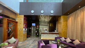 Un vestíbulo de hotel con una recepción, sofás morados, una mesa de centro de cristal y un suelo de mármol.