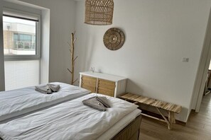 Ein Schlafzimmer mit zwei Betten, einer hölzernen Bank, einer weißen Kommode, einer gewebten Lampe und einem an der Wand hängenden Wandteller.