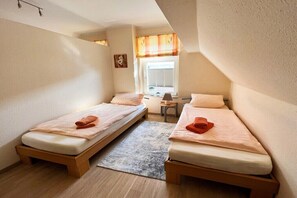 Ein kleines, gut beleuchtetes Zimmer mit zwei Einzelbetten, einem kleinen Tisch und einem Fenster mit Vorhängen.