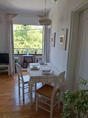 A dining area with a table set for four, white chairs, a hanging light fixture, and a view of greenery outside.