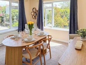 A dining area with a round table set for four, surrounded by chairs, with a view of greenery through large windows. - 2 bedroom accommodation in Porthleven (Porthleven)