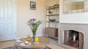 Un coin repas avec une table ronde, deux assiettes de nourriture, deux verres de jus d’orange, un vase avec des fleurs, un buffet en bois, une cheminée et un tableau encadré au mur.