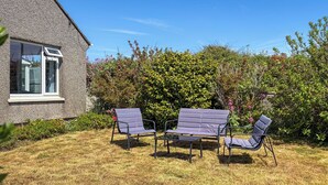 Une terrasse avec deux chaises et une table, entourée d’une haie et d’une maison.