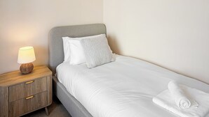 Un lit bien fait, avec une literie blanche, une tête de lit grise et une lampe de chevet posée sur une table de nuit en bois.