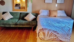 Ett rum med en grön soffa, en säng med ett mönstrat överkast, en väggpanel i trä och en liten köksdel med en mikrovågsugn.