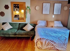 A room with a green sofa, a bed with a patterned bedspread, a wooden panel wall, and a small kitchen area with a microwave. - Stellar Ocean View, Amazing location!! (Honolulu)