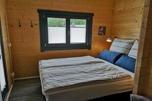 A wooden room with a bed, a window with blinds, a wall-mounted light, and a small picture.
