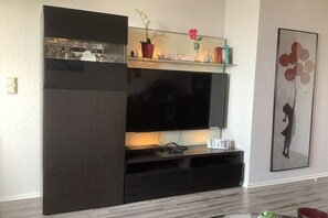 Ein modernes Wohnzimmer mit großem Flachbildfernseher, dunklen Holzmöbeln, einem Glastischchen mit Dekoration und einem Wandbild.