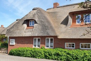 Ein traditionelles Haus mit Strohdach, Backsteinwänden, einem kleinen Fenster und einer Hecke davor.