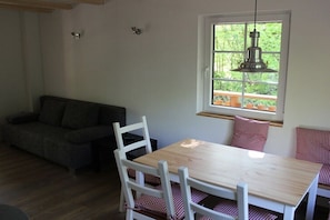 Ein Zimmer mit einem Holztisch, weißen Stühlen, einem grauen Sofa, einem Fenster mit Blick und einer hängenden Leuchte.