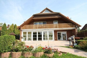 Ein zweistöckiges Haus mit einem Holzbalkon, großen Fenstern und einem Garten mit verschiedenen Pflanzen.