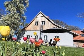 Ein Haus mit Holzfassade, einer weißen Wand und einem roten Dach. Vor dem Eingang stehen Stühle und eine Leiter.